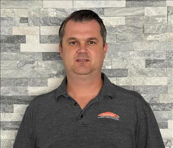 Male employee with short brown hair hair smiling in front of a grey brick wall.